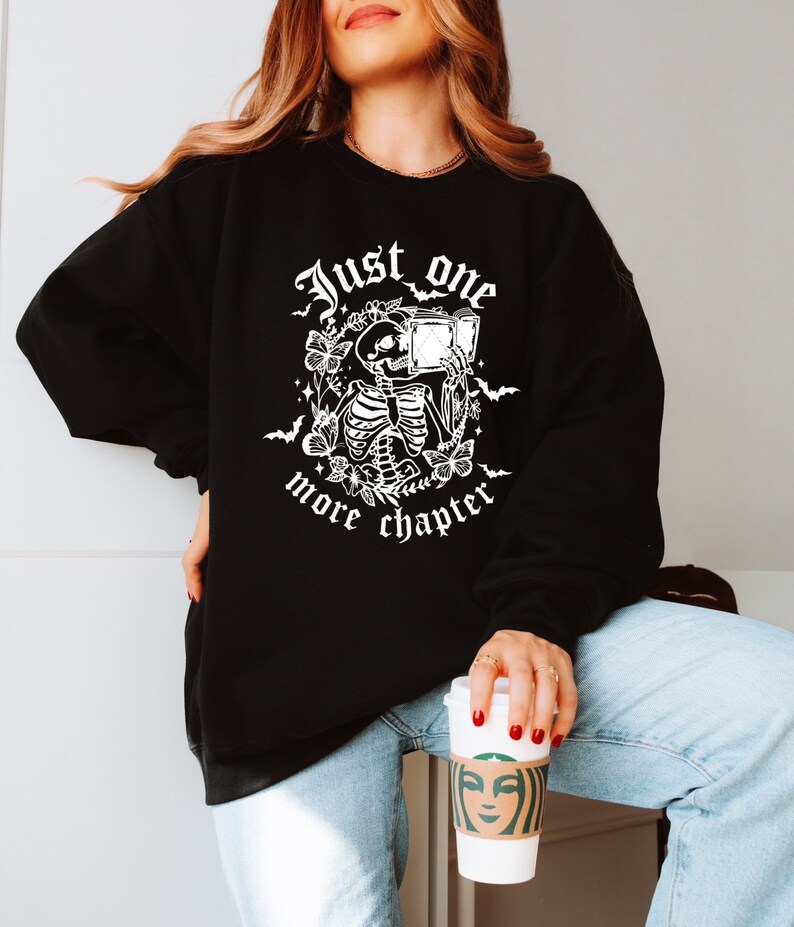 May include: Black sweatshirt with a white graphic of a skeleton reading a book, surrounded by flowers and bats. The text "Just one more chapter" is printed below the graphic. The sweatshirt is a casual style.