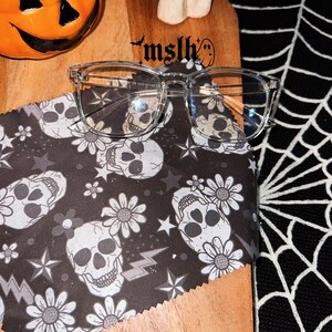 May include: Clear frame glasses with a skull and flower pattern on the cleaning cloth. The cloth is on a wooden surface with the text "mslh" on it.