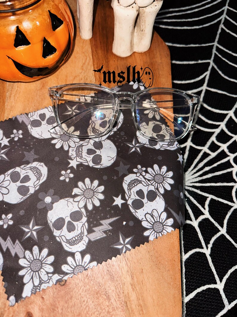 May include: Clear frame glasses with a skull and flower pattern on the cleaning cloth. The cloth is on a wooden surface with the text "mslh" on it.