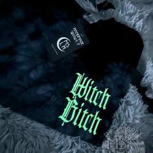 Puede incluir: Camiseta negra tie-dye con un gr&aacute;fico verde que brilla en la oscuridad que dice "Witch Bitch" en una fuente g&oacute;tica. La camiseta tambi&eacute;n tiene un peque&ntilde;o logotipo que dice "My Spooky Little Heart" en blanco.