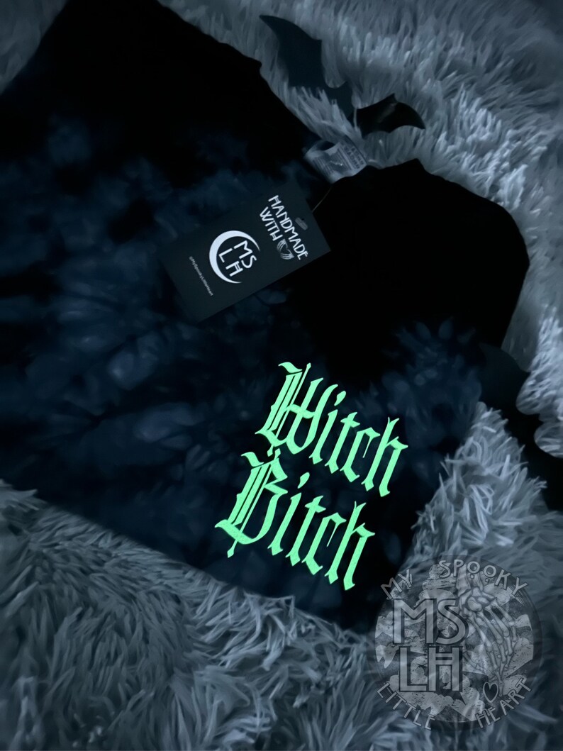 Puede incluir: Camiseta negra tie-dye con un gr&aacute;fico verde que brilla en la oscuridad que dice "Witch Bitch" en una fuente g&oacute;tica. La camiseta tambi&eacute;n tiene un peque&ntilde;o logotipo que dice "My Spooky Little Heart" en blanco.