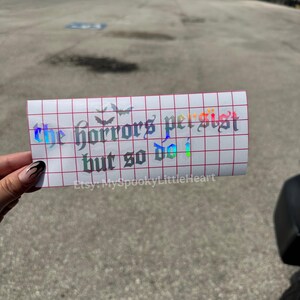May include: A rectangular holographic vinyl decal on a white grid transfer paper featuring the text "the horrors persist but so do i" in a gothic font, accompanied by small bat silhouettes and the text "Etsy: MySpookyLittleHeart".
