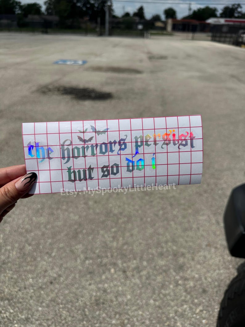 May include: A rectangular holographic vinyl decal on a white grid transfer paper featuring the text "the horrors persist but so do i" in a gothic font, accompanied by small bat silhouettes and the text "Etsy: MySpookyLittleHeart".