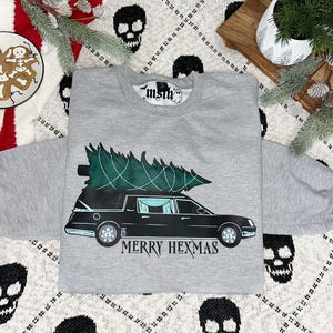 May include: A gray sweatshirt with a black hearse carrying a Christmas tree on top. The text "Merry Hexmas" is printed below the hearse.