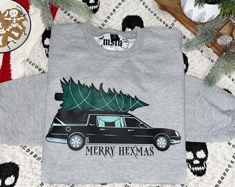 May include: A gray sweatshirt with a black hearse carrying a Christmas tree on top. The text "Merry Hexmas" is printed below the hearse.