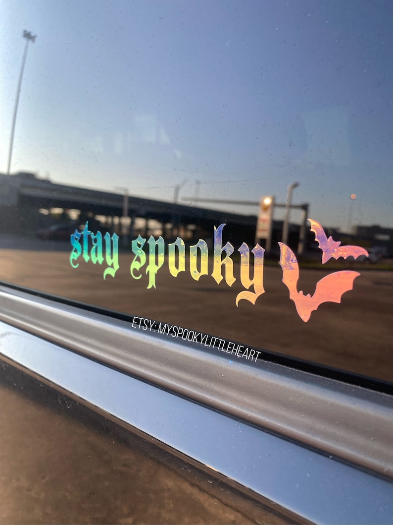 Puede incluir: Una pegatina hologr&aacute;fica en la ventana de un coche con la frase "stay spooky" en una fuente g&oacute;tica. Dos siluetas de murci&eacute;lagos est&aacute;n a la derecha del texto. El nombre de la tienda Etsy "myspookylittleheart" est&aacute; debajo.