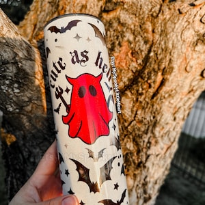 May include: A white tumbler with a black and red ghost design. The tumbler has the text "cute as hell" on it and is surrounded by black bats.