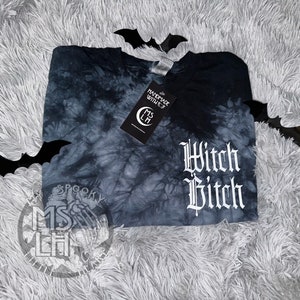 Puede incluir: Una camiseta negra tie-dye con el texto "Witch Bitch" en letras g&oacute;ticas blancas. La camiseta est&aacute; sobre una superficie blanca y peluda con murci&eacute;lagos negros.