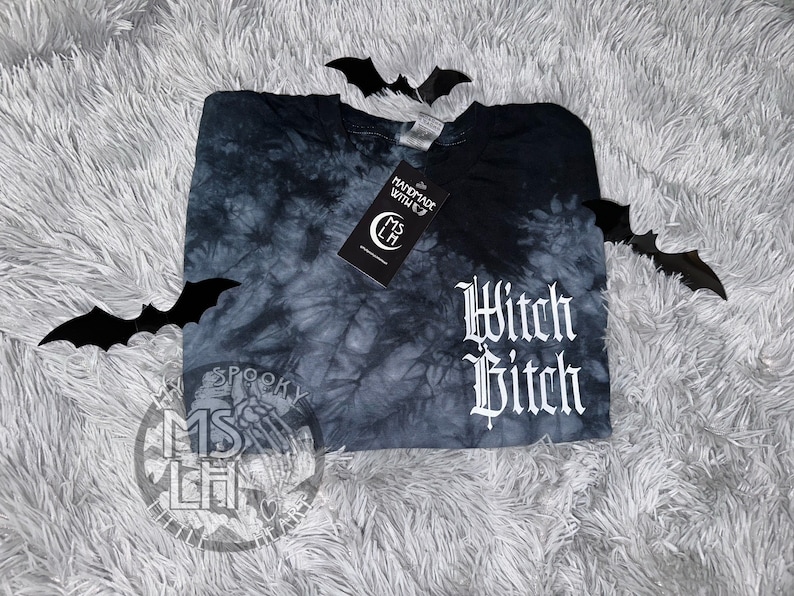 Puede incluir: Una camiseta negra tie-dye con el texto "Witch Bitch" en letras g&oacute;ticas blancas. La camiseta est&aacute; sobre una superficie blanca y peluda con murci&eacute;lagos negros.