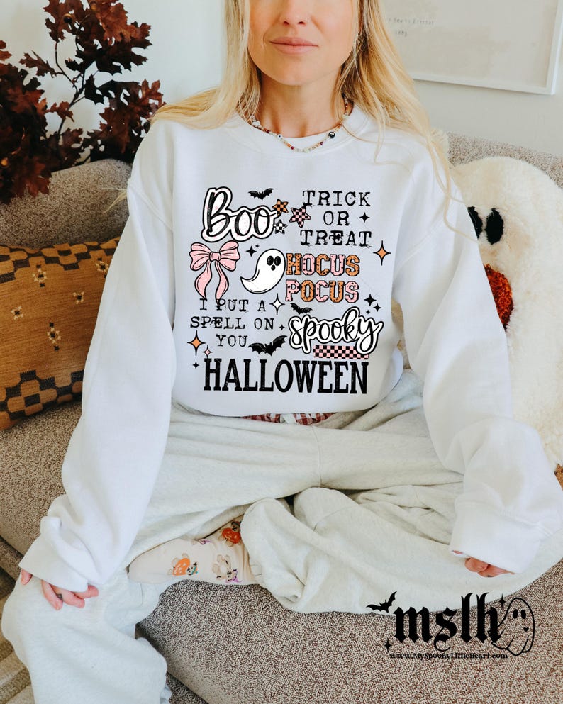 May include: White Halloween sweatshirt with the text "Boo Trick or Treat Hocus Pocus Spooky Halloween". The design includes a ghost, bow, and bat graphics. The sweatshirt is part of a Halloween-themed collection.