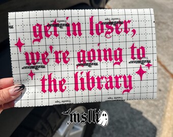 May include: A rectangular sheet of white transfer tape with a grid pattern featuring the hot pink text get in loser, we're going to the library in a gothic font style, accented by four small four-pointed star shapes on the sides of the lettering.