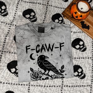 May include: A gray t-shirt with a distressed design featuring a black raven perched on roses, the text "F-CAW-F", a crescent moon, and stars. The shirt is on a gray skull-patterned blanket, with a pumpkin candle holder and wooden tray.