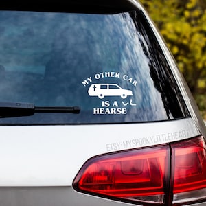 May include: White vinyl sticker with the text "My other car is a hearse" and a cartoon image of a hearse with a cross on the roof. There are also two bats flying above the hearse. The sticker is on the back window of a silver car.