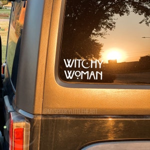 May include: A white vinyl sticker on a brown car window that says "Witchy Woman" with a crescent moon and star symbol.