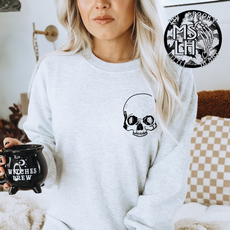 May include: A gray sweatshirt with a black skull graphic on the front. The sweatshirt has a crew neck and long sleeves. The model is holding a black mug that says "Witches Brew".