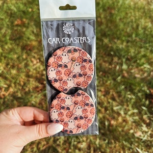 May include: Two round car coasters with a Halloween theme. The coasters feature a pink background with a repeating pattern of ghosts, pumpkins, bats, and candy corn.
