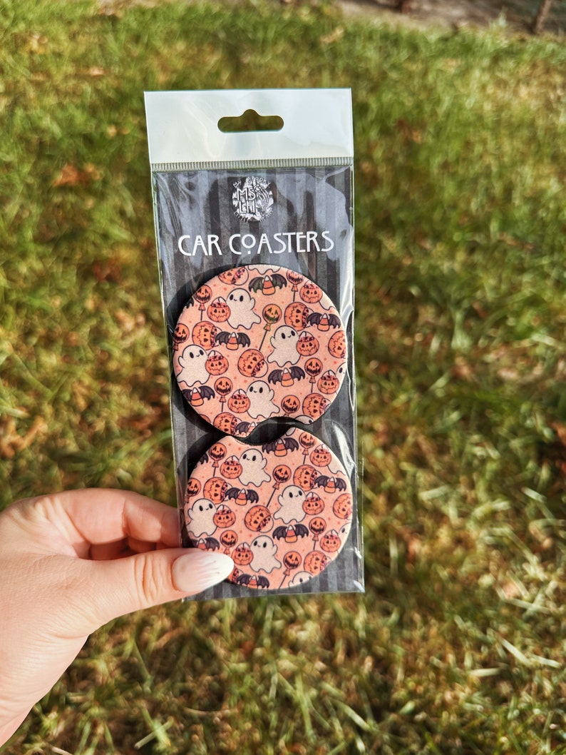 May include: Two round car coasters with a Halloween theme. The coasters feature a pink background with a repeating pattern of ghosts, pumpkins, bats, and candy corn.