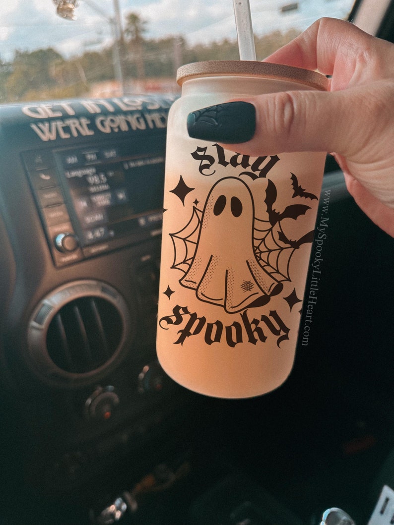 May include: A white tumbler with a black and white ghost graphic and the text "slay spooky" and a website address. The tumbler has a black lid and a straw.