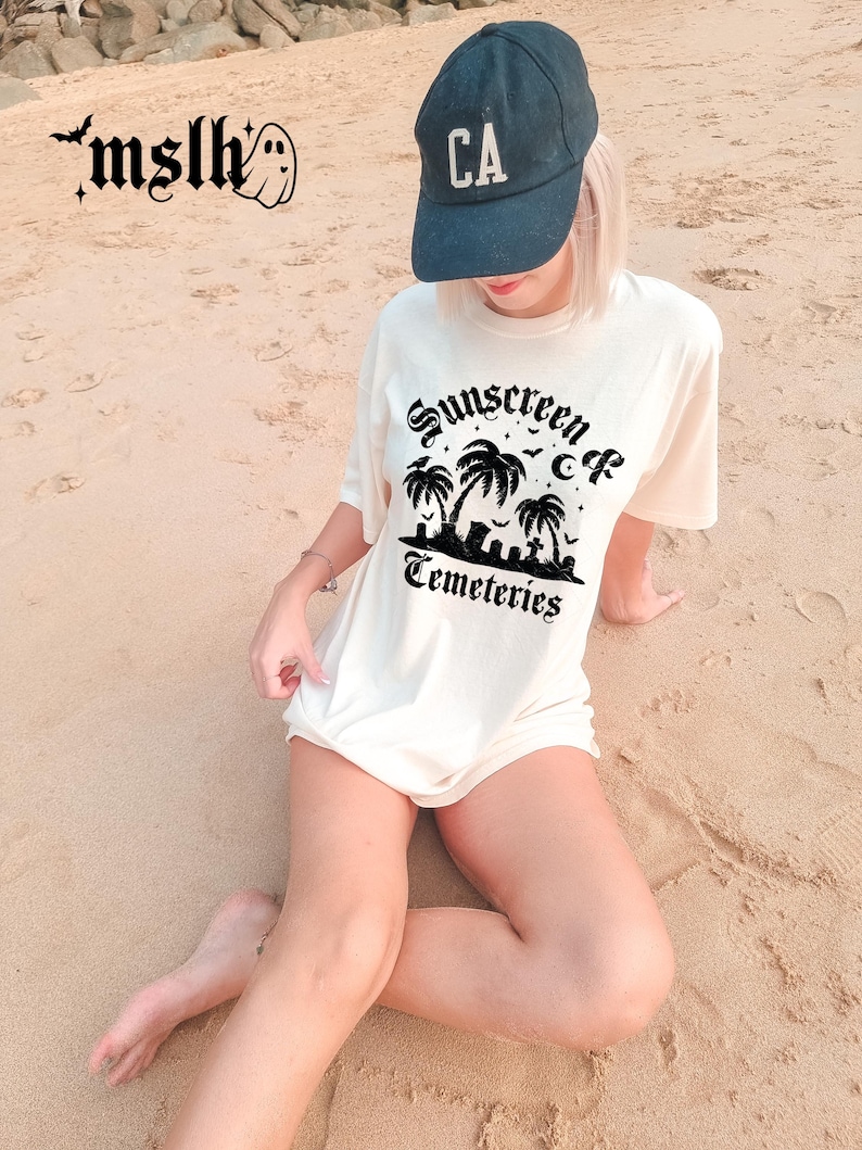 May include: Cream-colored t-shirt with the text "Sunscreen & Cemeteries" in a gothic font, featuring a palm tree and cemetery graphic. A dark blue baseball cap with "CA" is worn. The image is taken on a sandy beach.