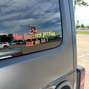 May include: A vinyl decal applied to a vehicle window featuring the text "the horrors persist but so do i" in a gothic-style font. The lettering displays a vibrant gradient of red, orange, and yellow, accented by three small bat silhouettes.