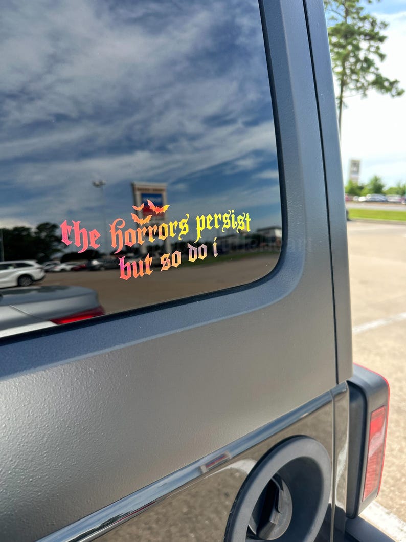 May include: A vinyl decal applied to a vehicle window featuring the text "the horrors persist but so do i" in a gothic-style font. The lettering displays a vibrant gradient of red, orange, and yellow, accented by three small bat silhouettes.
