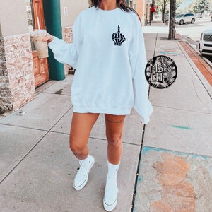 May include: White sweatshirt with a black skeleton hand giving the middle finger graphic on the front. The model is wearing white sneakers and holding a coffee cup.