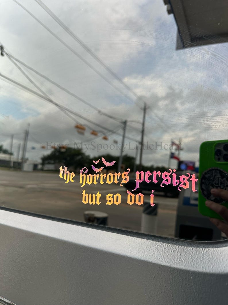 May include: A holographic vinyl decal applied to a window, featuring the text "the horrors persist but so do i" in a gothic-style font. The design includes three small bat silhouettes above the text, with a color-shifting gradient effect.