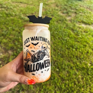May include: A frosted glass tumbler with a bamboo lid and a clear straw featuring a black bat-shaped straw topper. The glass displays a black graphic of a skeleton reclining with the text "JUST WAITING FOR HALLOWEEN" and small star and candy icons.
