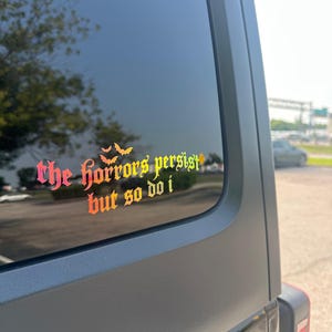 May include: A vinyl decal applied to a vehicle window featuring the text "the horrors persist but so do i" in a gothic-style font with a gradient color scheme transitioning from pink to orange and yellow, accented by three small bat silhouettes.