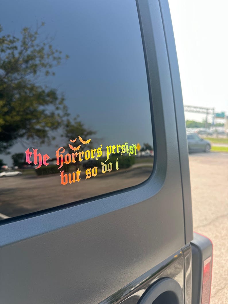 May include: A vinyl decal applied to a vehicle window featuring the text "the horrors persist but so do i" in a gothic-style font with a gradient color scheme transitioning from pink to orange and yellow, accented by three small bat silhouettes.