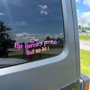 May include: A pink and purple gradient vinyl decal applied to a dark vehicle window. The decal features the text "the horrors persist but so do i" in a gothic-style font, accompanied by a small graphic of three bats above the word horrors.