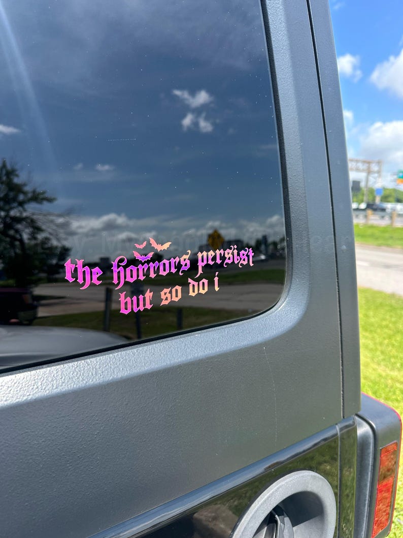 May include: A pink and purple gradient vinyl decal applied to a dark vehicle window. The decal features the text "the horrors persist but so do i" in a gothic-style font, accompanied by a small graphic of three bats above the word horrors.