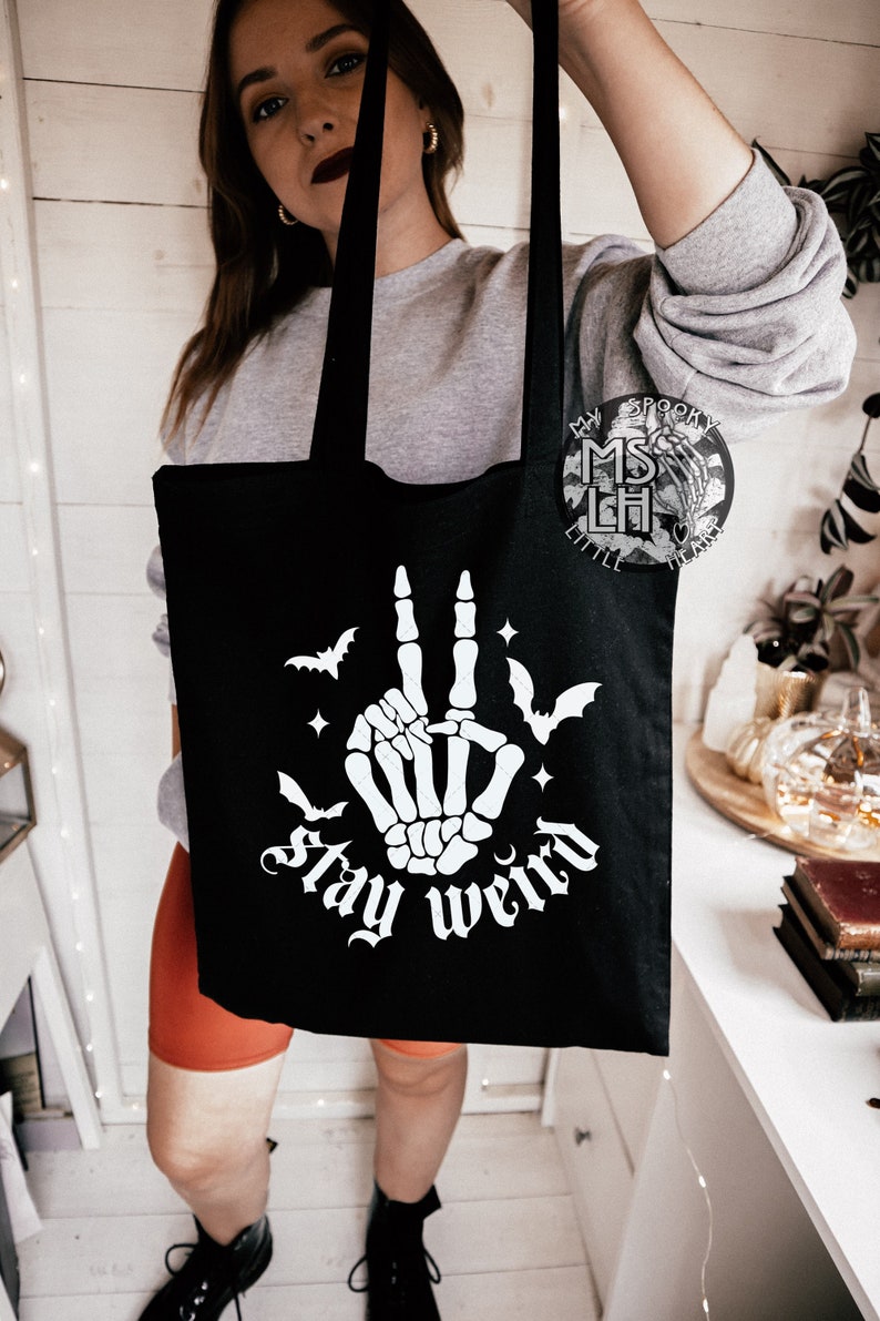 May include: Black tote bag with a white skeleton hand graphic making a peace sign, bats, stars, and the words "stay weird". The bag has long black handles.