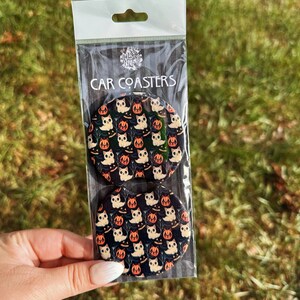 May include: Two round black car coasters with a white cat and orange pumpkin pattern. The coasters are packaged in a clear plastic bag with a white header that says "CAR COASTERS".