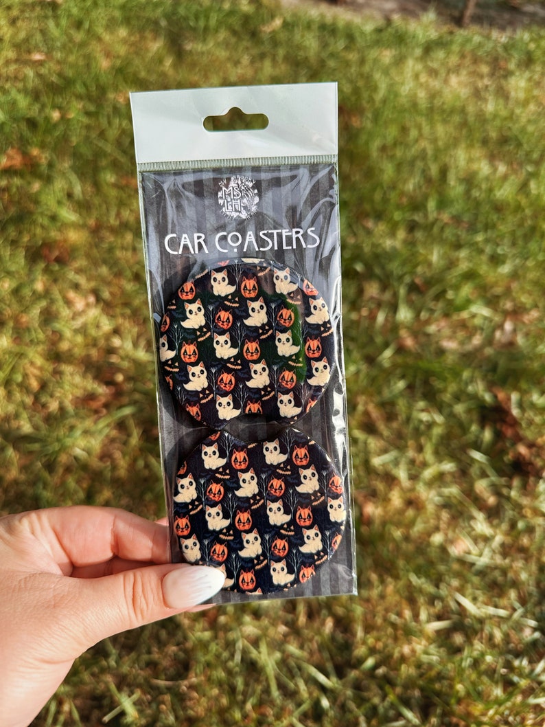 May include: Two round black car coasters with a white cat and orange pumpkin pattern. The coasters are packaged in a clear plastic bag with a white header that says "CAR COASTERS".
