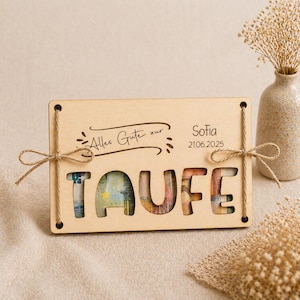 Könnte beinhalten: Eine hellfarbene Holzgeschenkbox mit dem ausgeschnittenen Wort "TAUFE", das Geld im Inneren zeigt. Die Box trägt den Text "Alles Gute zur" und "Sofia 21.06.2025". Sie ist mit Bindfäden gebunden und hat eine dekorative Schleife.