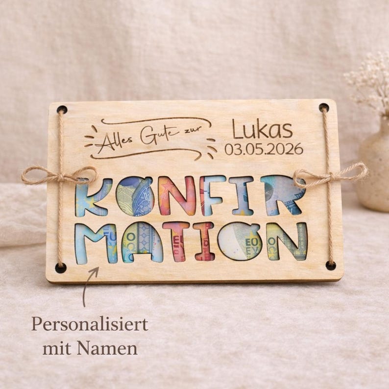 May include: A wooden plaque with the word "KONFIRMATION" cut out, revealing colorful money inside. The plaque has the name "Lukas" and the date "03.05.2026" engraved. It is tied with twine and has the text "Personalisiert mit Namen".