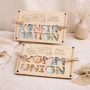 May include: Two wooden plaques with cut-out letters spelling "KONFIRMATION" and "KOMMUNION", filled with Euro banknotes. Each plaque has a name and date, tied with twine. The background includes a vase with flowers.