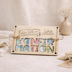May include: Wooden plaque with the word "KONFIRMATION" cut out, filled with colorful currency. The plaque has the text "Alles Gute zur Lukas 03.05.2026" and is tied with twine. A decorative gift for a confirmation.