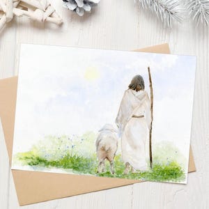 May include: Watercolor art print of a shepherd with a sheep, walking on a path. The shepherd is wearing a white robe and holding a wooden staff. The sheep is white. The background is a light blue sky with a yellow sun. The print is on a white card with a brown envelope.