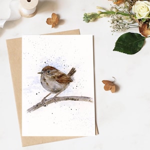 May include: A watercolor painting of a small brown bird perched on a branch, set against a white background with blue and black speckles. The artwork is on a white card, placed on a brown envelope. Dried flowers and a ribbon are in the background.