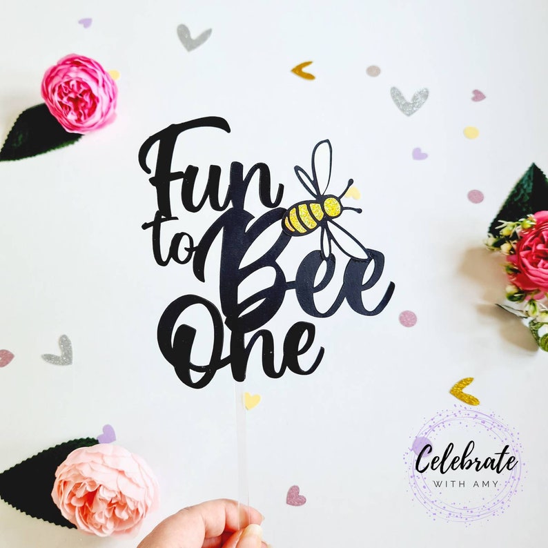 May include: Black cake topper with the text "Fun to Bee One" and a yellow and black bee with gold glitter accents.
