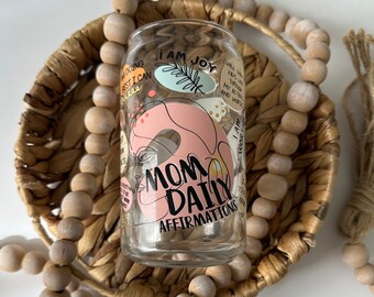 May include: A clear glass cup with a pink and blue design and the text "Mom Daily Affirmations" printed on it. The cup is sitting in a woven basket with wooden beads.