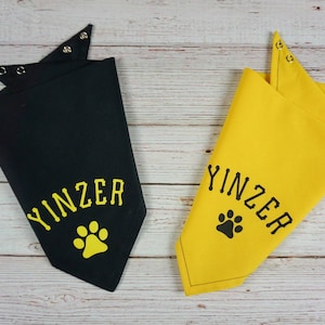 May include: Two dog bandanas, one black and one yellow, with the word "YINZER" and a paw print. The bandanas are folded and have metal snap closures at the top. The background is a light wood surface.