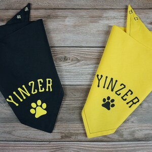 May include: Two dog bandanas, one black and one yellow, with the word "YINZER" and a paw print design. The bandanas have snap closures at the neck and are displayed on a wooden surface.