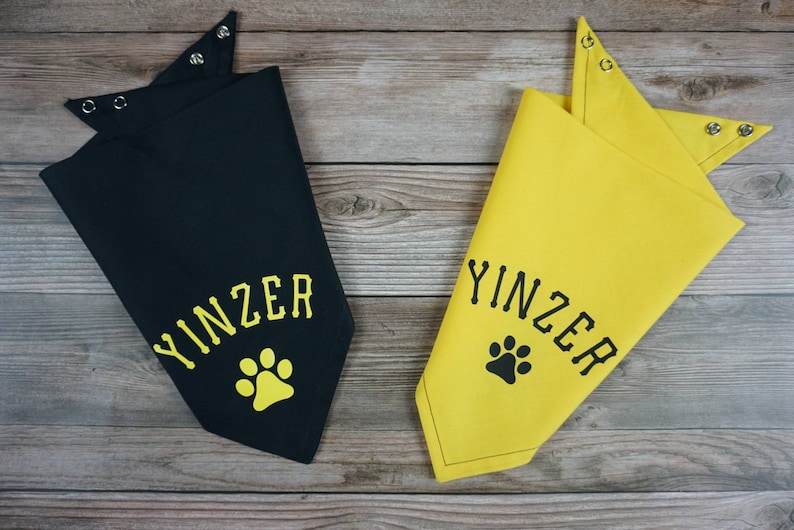 May include: Two dog bandanas, one black and one yellow, with the word "YINZER" and a paw print design. The bandanas have snap closures at the neck and are displayed on a wooden surface.