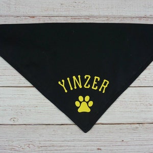 May include: Black triangular dog bandana with the word "YINZER" in yellow lettering and a yellow paw print. The bandana has metal grommets at the corners for easy tying. A pet accessory.