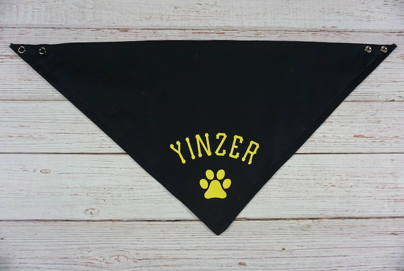 May include: Black triangular dog bandana with the word "YINZER" in yellow lettering and a yellow paw print. The bandana has metal grommets at the corners for easy tying. A pet accessory.