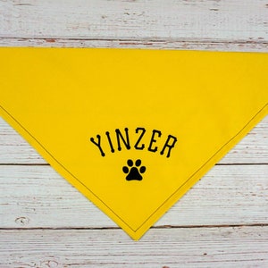 May include: A bright yellow dog bandana with the word "YINZER" in black block letters and a black paw print. The bandana is triangular, with metal grommets at the corners. The item is made of fabric.
