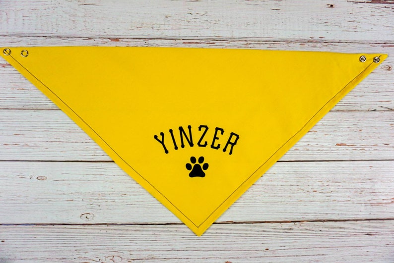 May include: A bright yellow dog bandana with the word "YINZER" in black block letters and a black paw print. The bandana is triangular, with metal grommets at the corners. The item is made of fabric.
